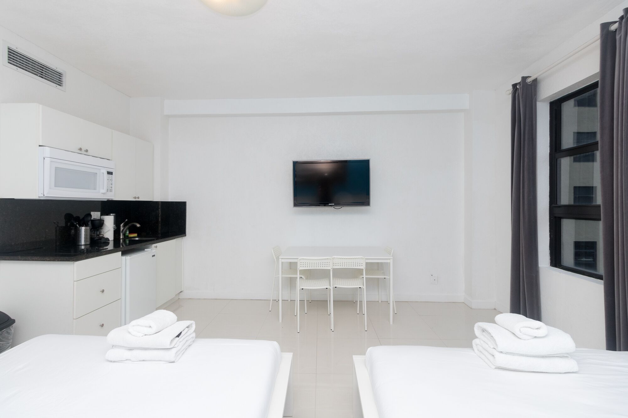 A modern hotel room with a kitchenette, a flat-screen TV, a small table, two beds, and a large window.