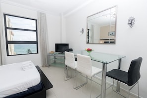Une chambre d’hôtel comprenant un lit, un bureau avec un ordinateur, deux chaises, une table en verre et une grande fenêtre donnant sur l’océan.