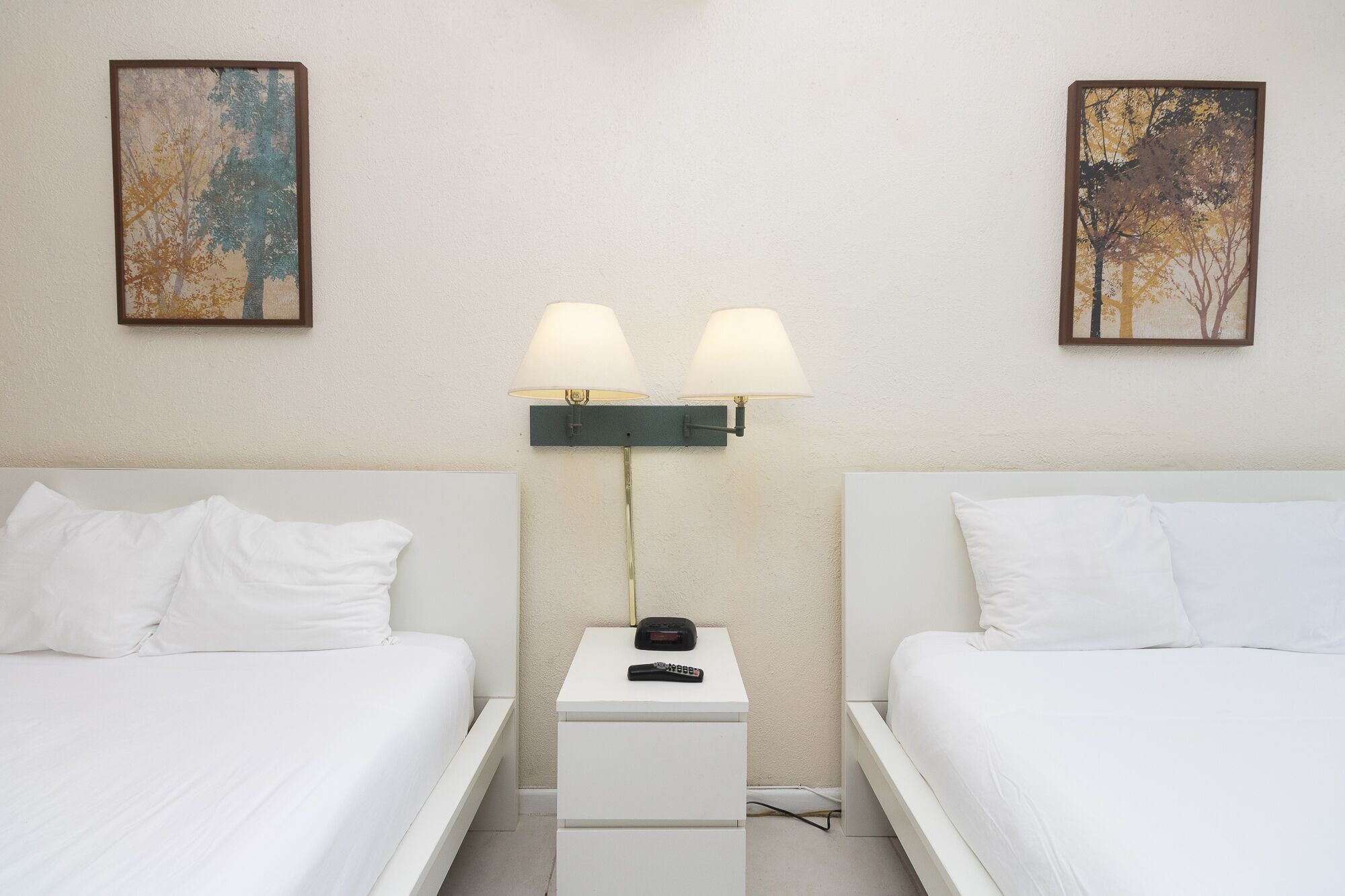 A hotel room with two single beds, a nightstand with a lamp, and two paintings on the wall.