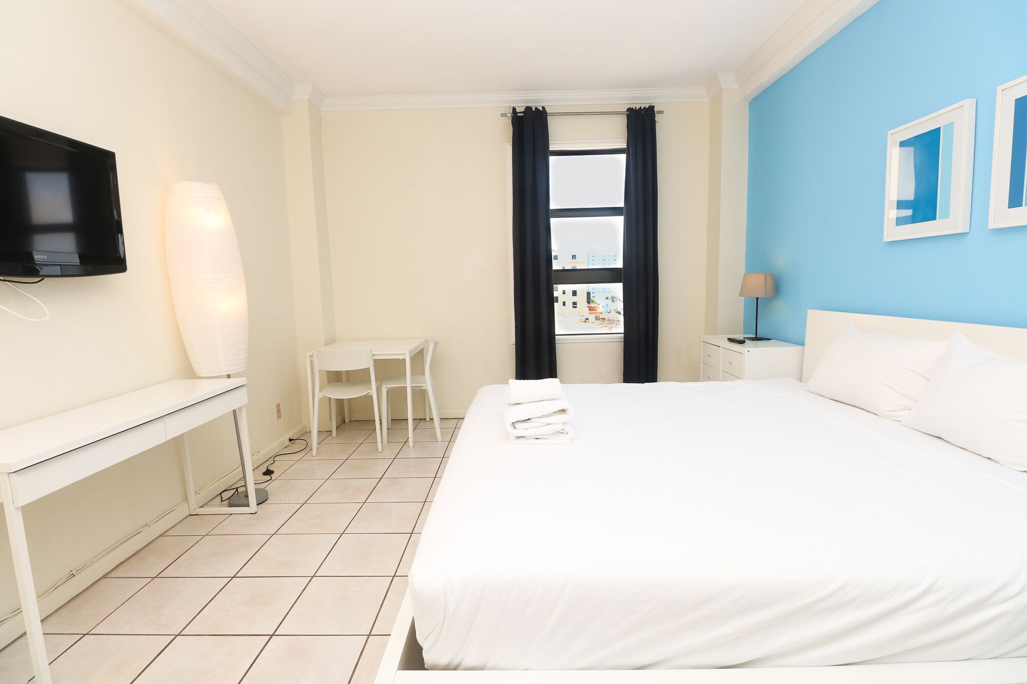 A hotel room with a bed, a desk, a chair, a TV, a window with curtains, and a lamp.