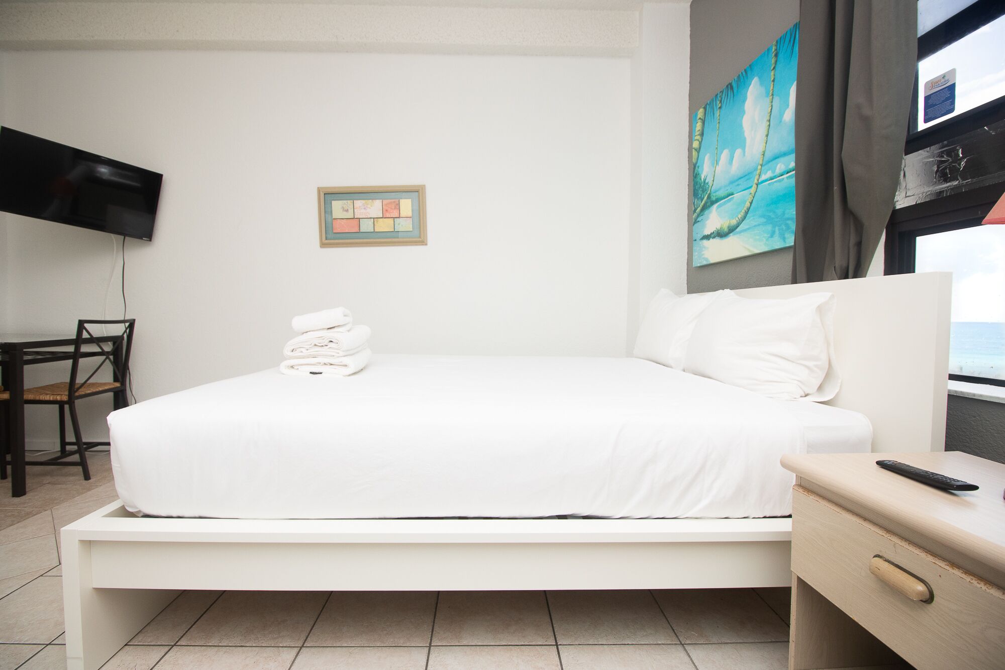 A neatly made bed with white linens, a small table with a remote control, a chair, and a painting on the wall.