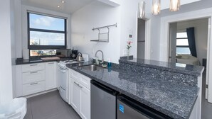 A modern kitchen with white cabinets, a granite countertop, a dishwasher, and a window overlooking a cityscape. - 701 Ocean View Hollywood Beach (Hollywood)