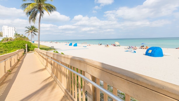 Een strand met een houten looppad, palmbomen en blauwe strandparasols.