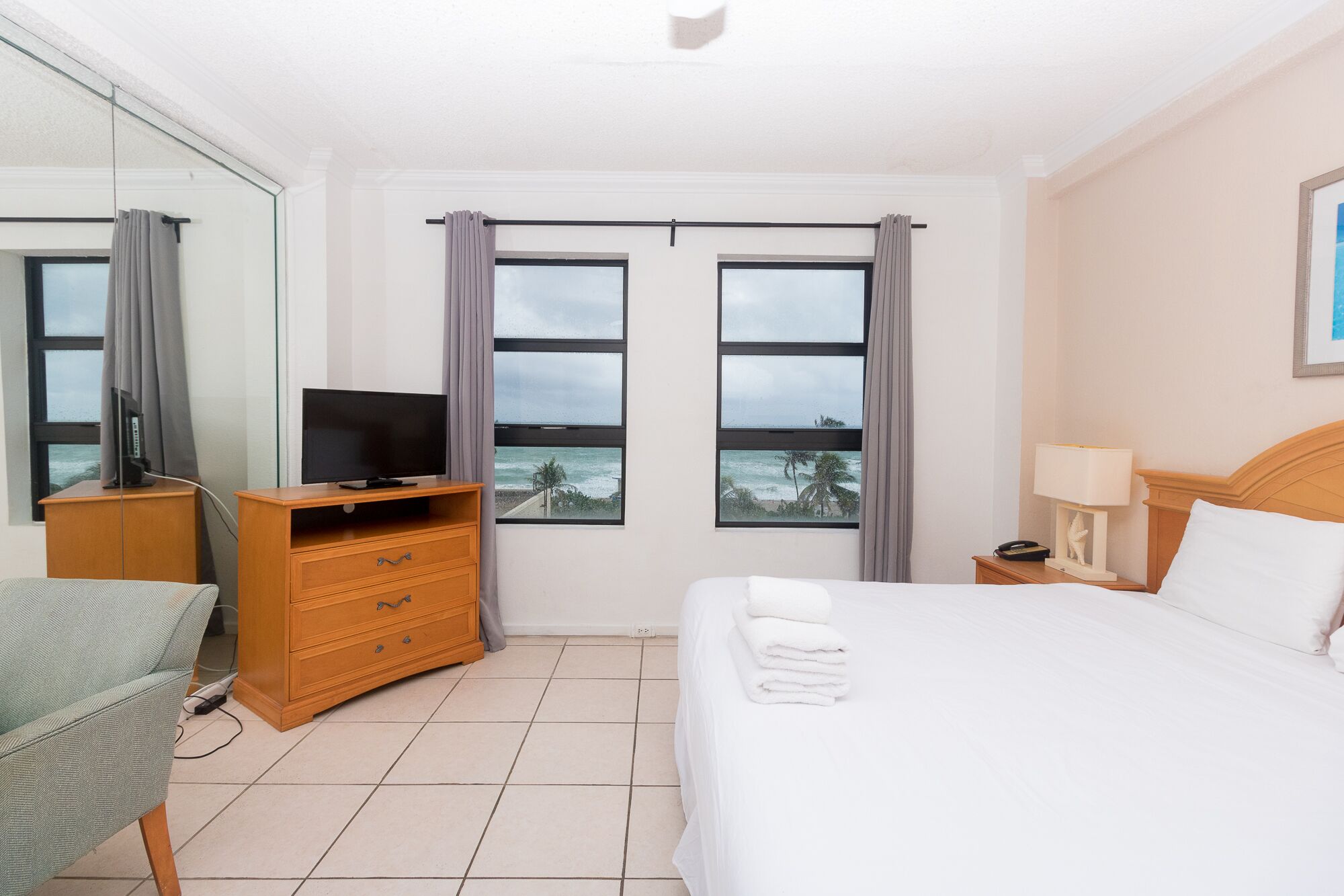 A hotel room with a bed, a TV on a wooden dresser, a lamp, and a view of the ocean through large windows.