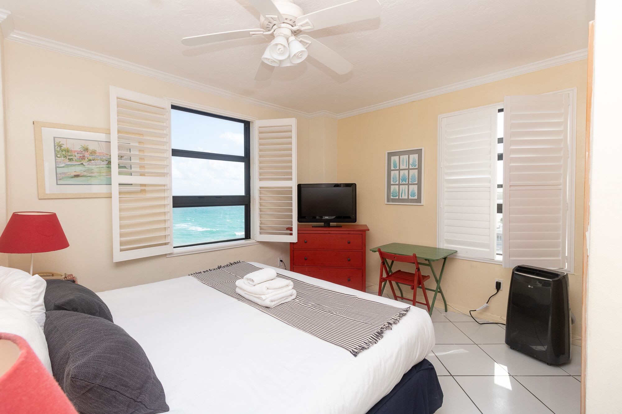 A hotel room with a bed, a TV, a desk, and a view of the ocean through the window.