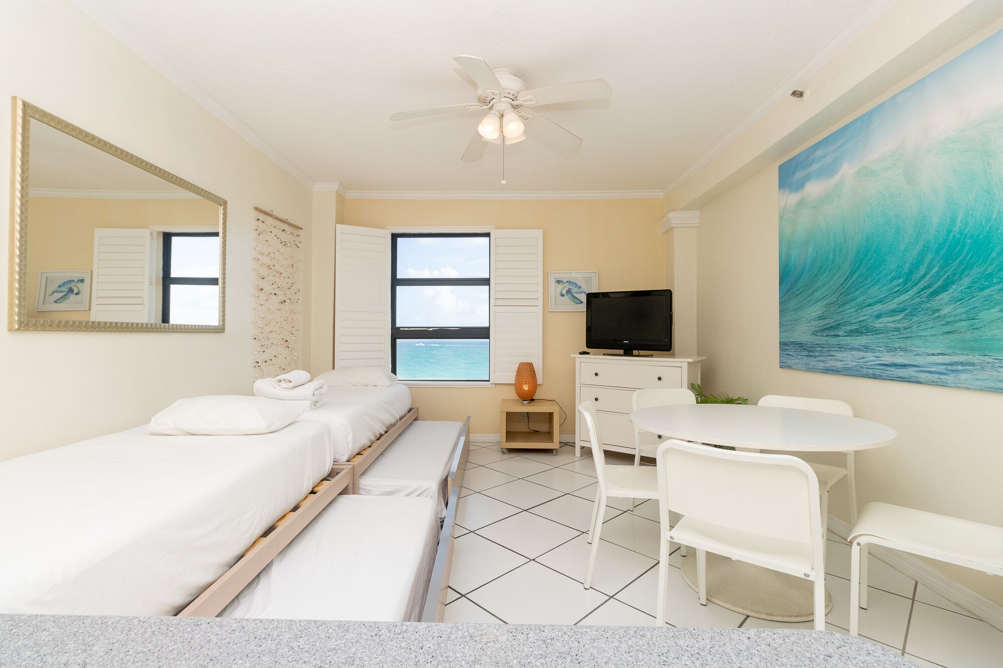 A hotel room with a bed, a desk, a chair, a television, a ceiling fan, a window with a view of the ocean, and a painting of a wave.