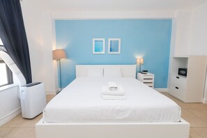 A neatly made bed with white linens and a folded towel, a bedside table with a lamp, and a small cabinet with a radio.