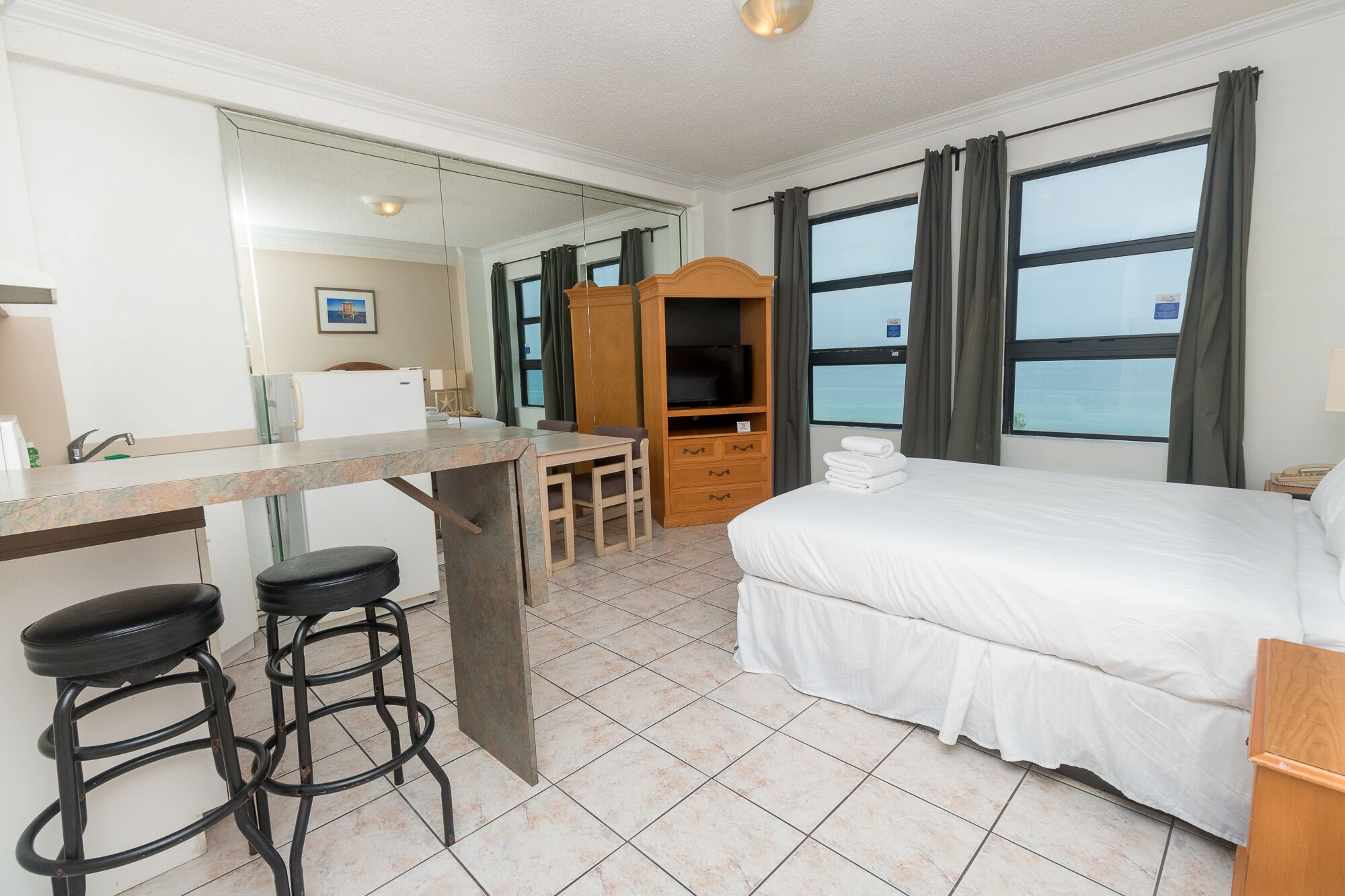 Ein Hotelzimmer mit einem Bett, einem Schreibtisch mit zwei Hocker, einem Fernseher und Meerblick durch große Fenster.