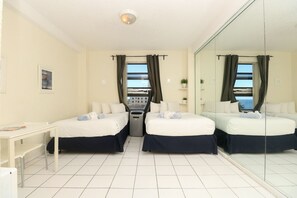 A hotel room with two beds, a desk, a chair, a small table, a mirror, and a view of the sea. - 588 Partial Ocean View (Hollywood)