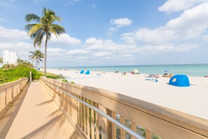 Een strand met een houten looppad, palmbomen en blauwe strandparasols.