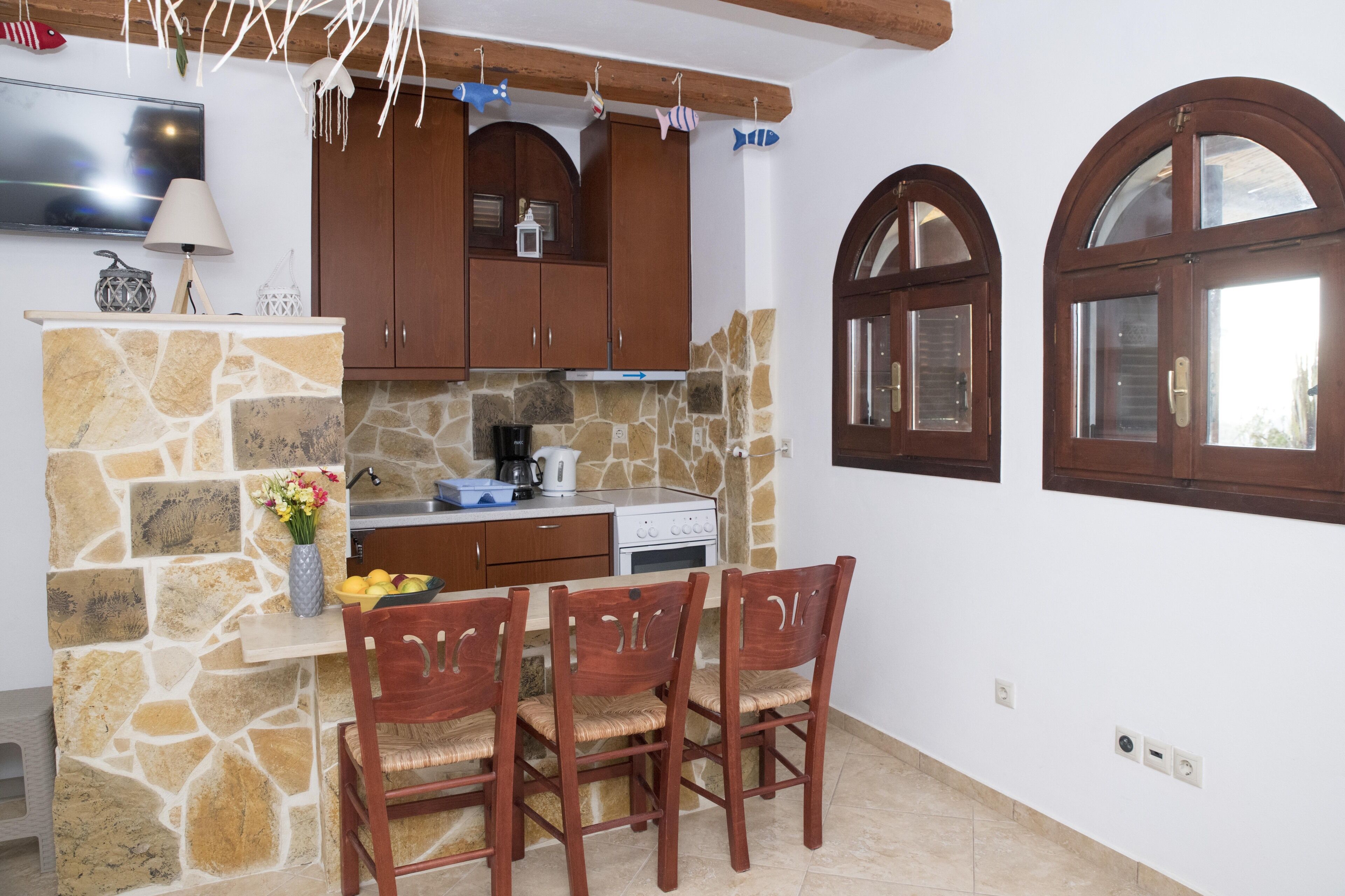 A kitchen with stone walls, wooden cabinets, a dining table with chairs, and arched windows.