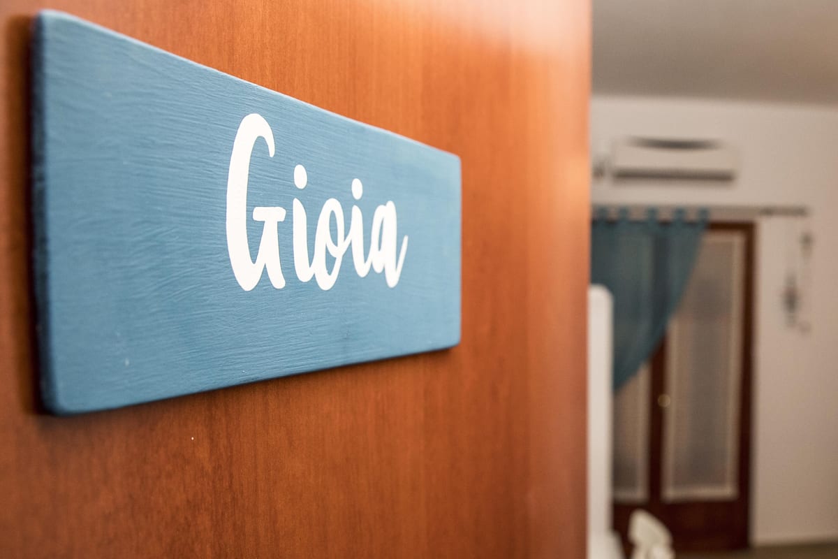 Une plaque bleue portant le nom « Gioia » est fixée à une porte en bois.