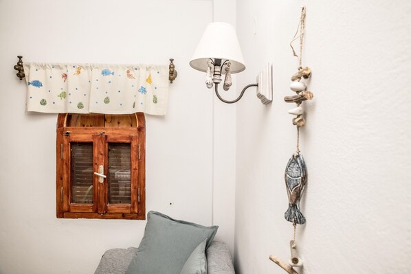 Una habitación con ventana de madera, un sofá gris, pared blanca, una lámpara y decoración en la pared.