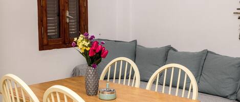 A dining area with a wooden table, white chairs, a vase with flowers, and a grey sofa.