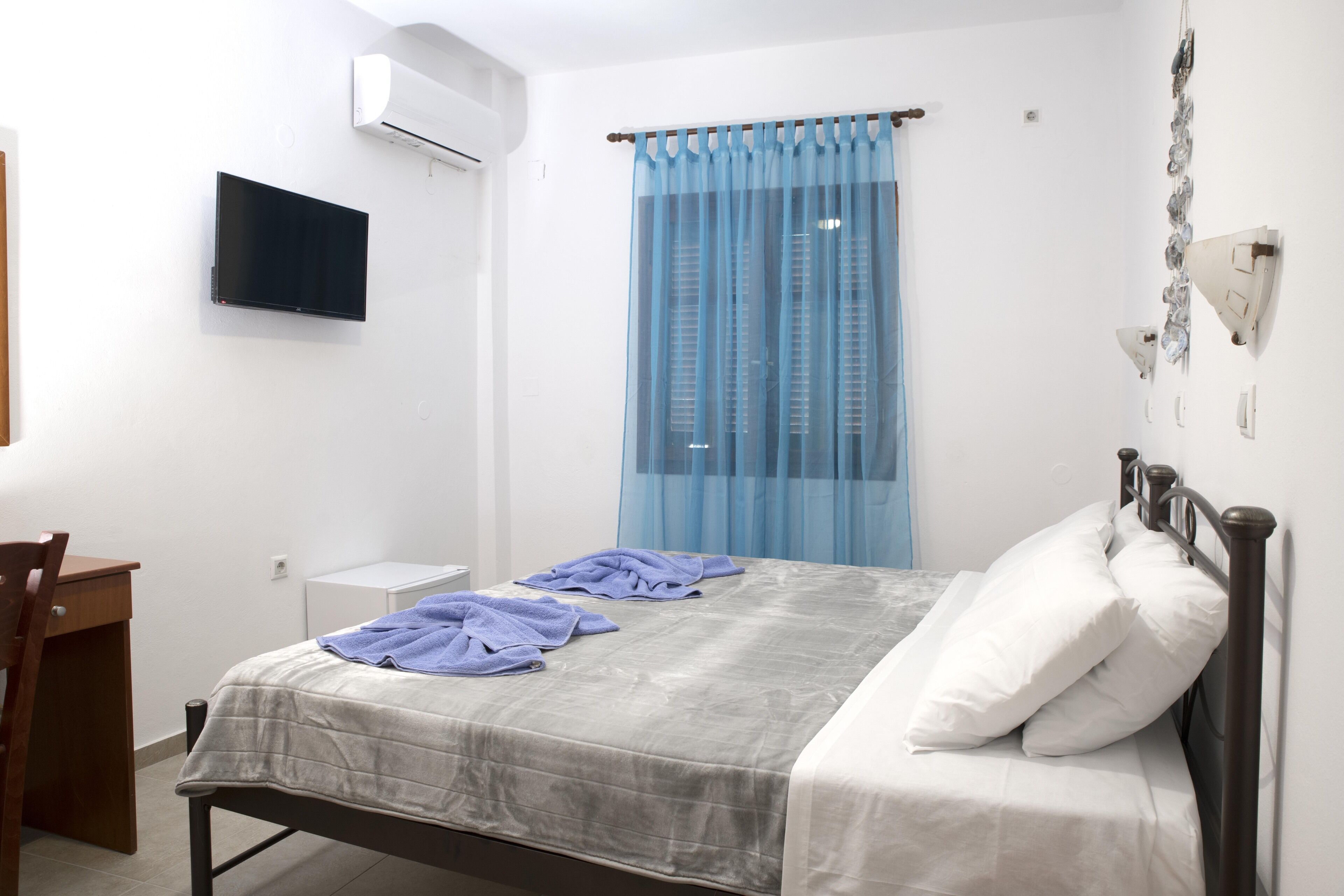 A bedroom with a bed, a desk, a chair, a TV, and a window with curtains.