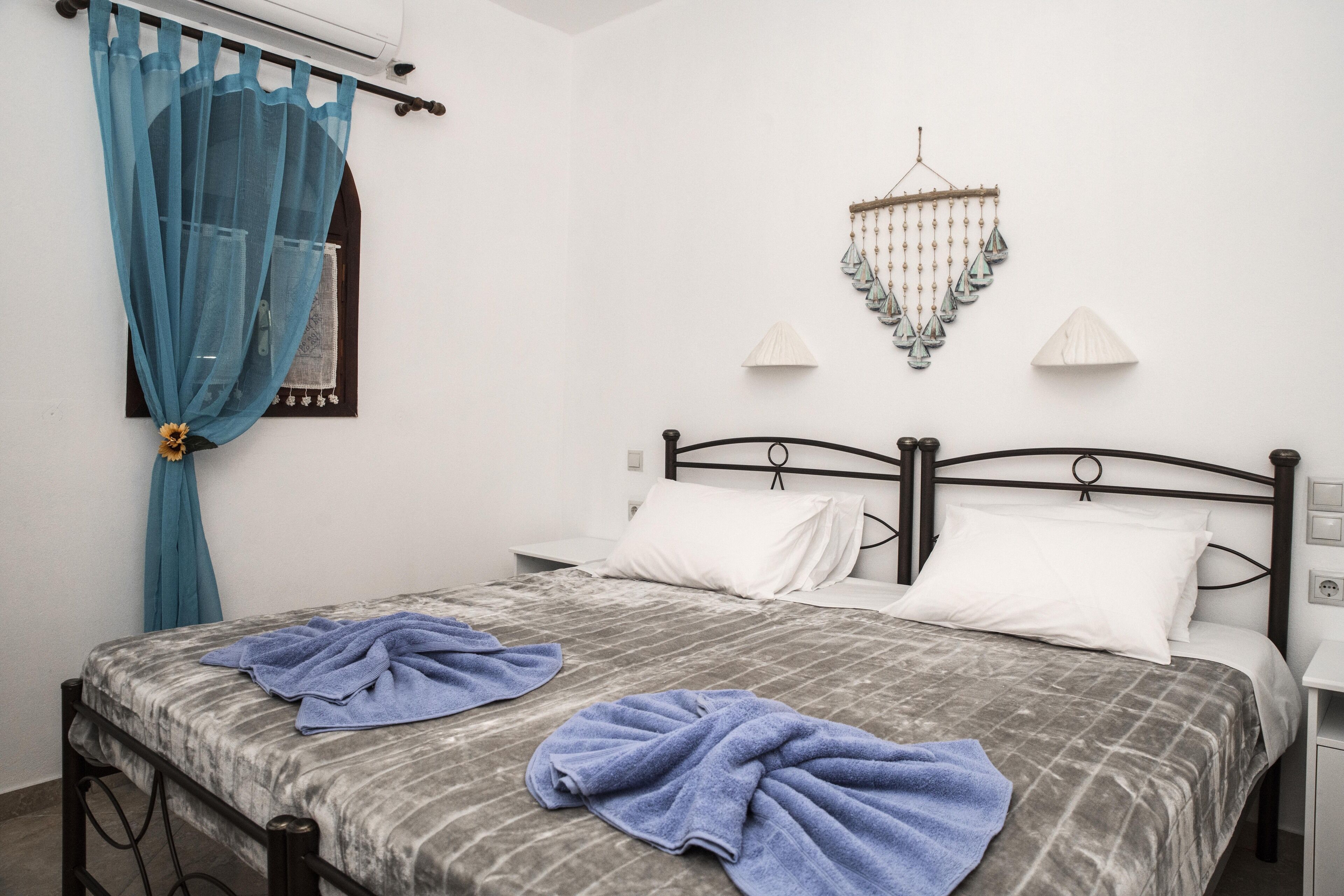 A double bed with a grey and white checkered bedspread, two white pillows, and two blue towels. There is a small bedside table with a lamp and a wall-mounted decorative item.