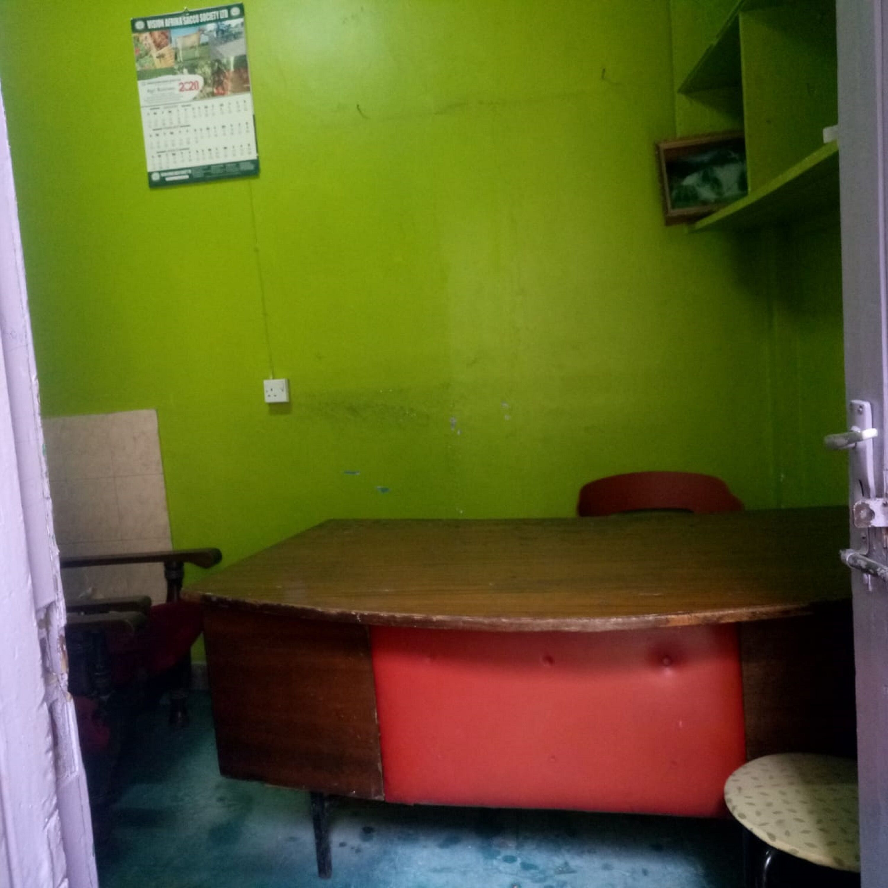 A room with a green wall, a wooden table with a red cushion, a chair, and a small shelf with a picture frame.