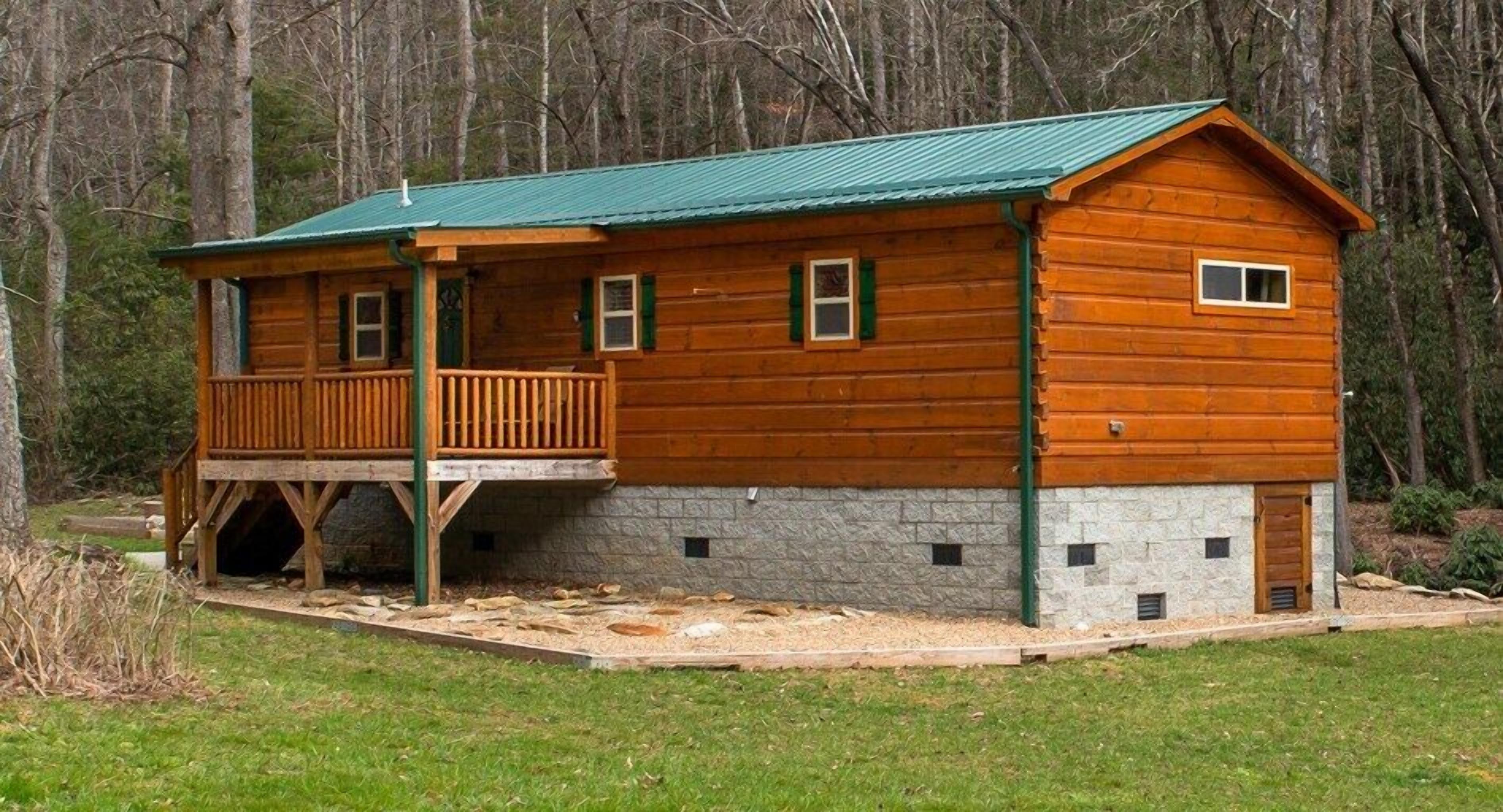 A log cabin with a porch, stone foundation, and a green roof.