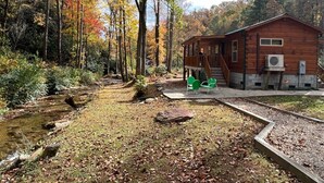 A cabin with a deck, a small stream, and a forested backdrop. - Creekside Log Cabin in Pisgah Forest (Pisgah Forest)