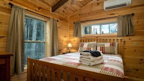 A wooden cabin bedroom with a bed, curtains, a window, and an air conditioning unit. - Creekside Log Cabin in Pisgah Forest (Pisgah Forest)