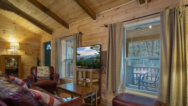 A wooden cabin interior with a television, leather furniture, and a balcony with outdoor seating. - Creekside Log Cabin in Pisgah Forest (Pisgah Forest)