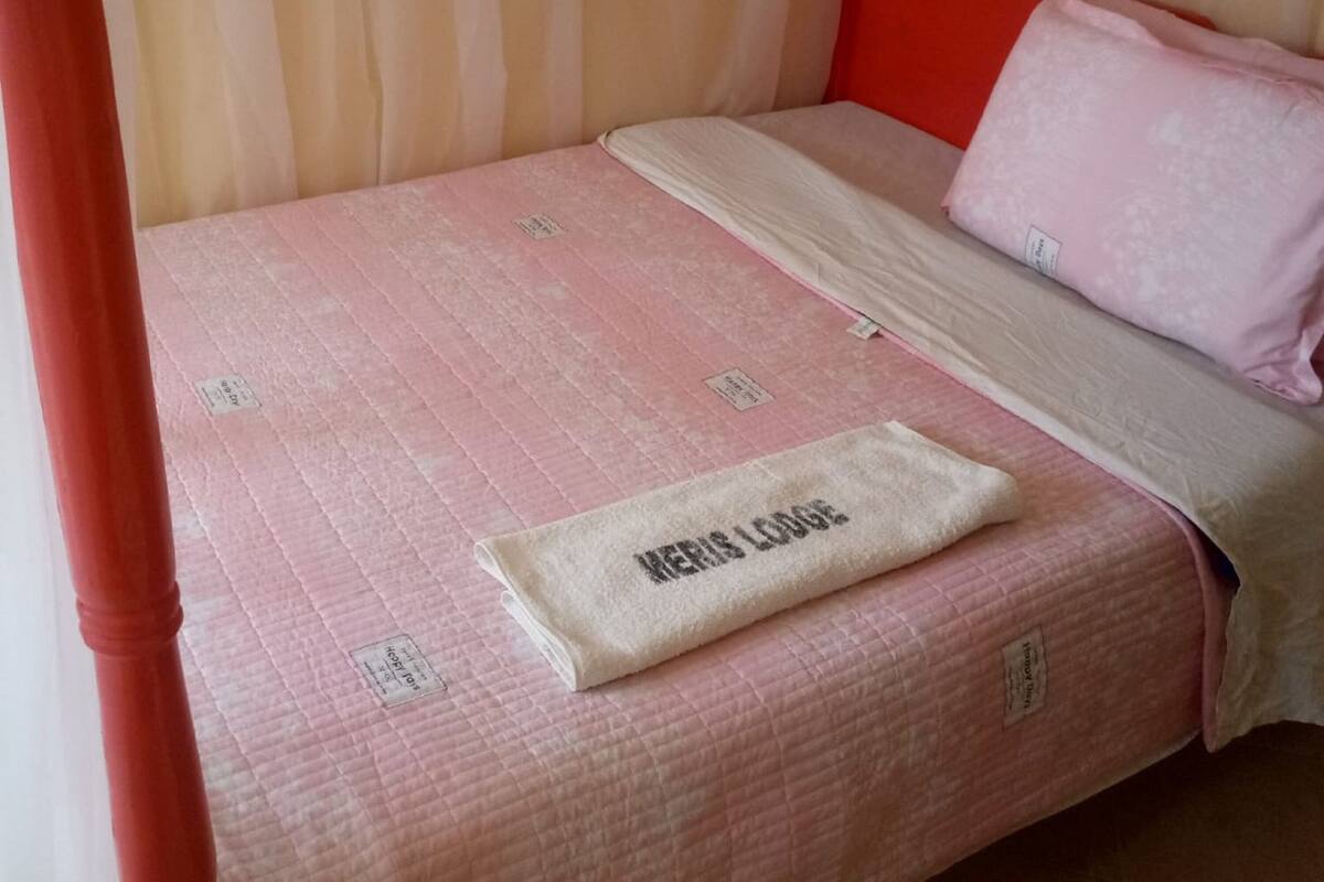 Una cama con colcha rosa, almohada blanca y manta color beige con una etiqueta.