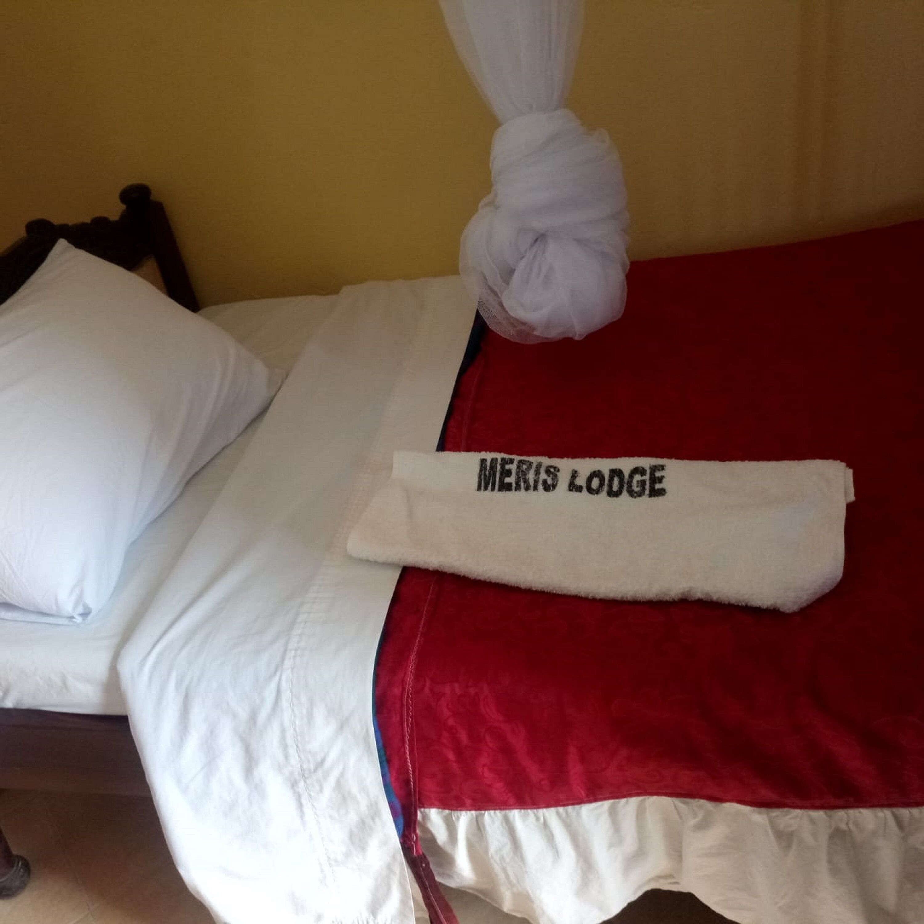 A bed with white pillows, a red blanket, and a towel with "MERIS LODGE" text.