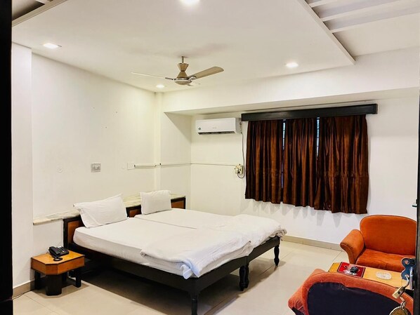 A hotel room with a bed, a nightstand, a telephone, an air conditioning unit, a window with curtains, and an orange chair.