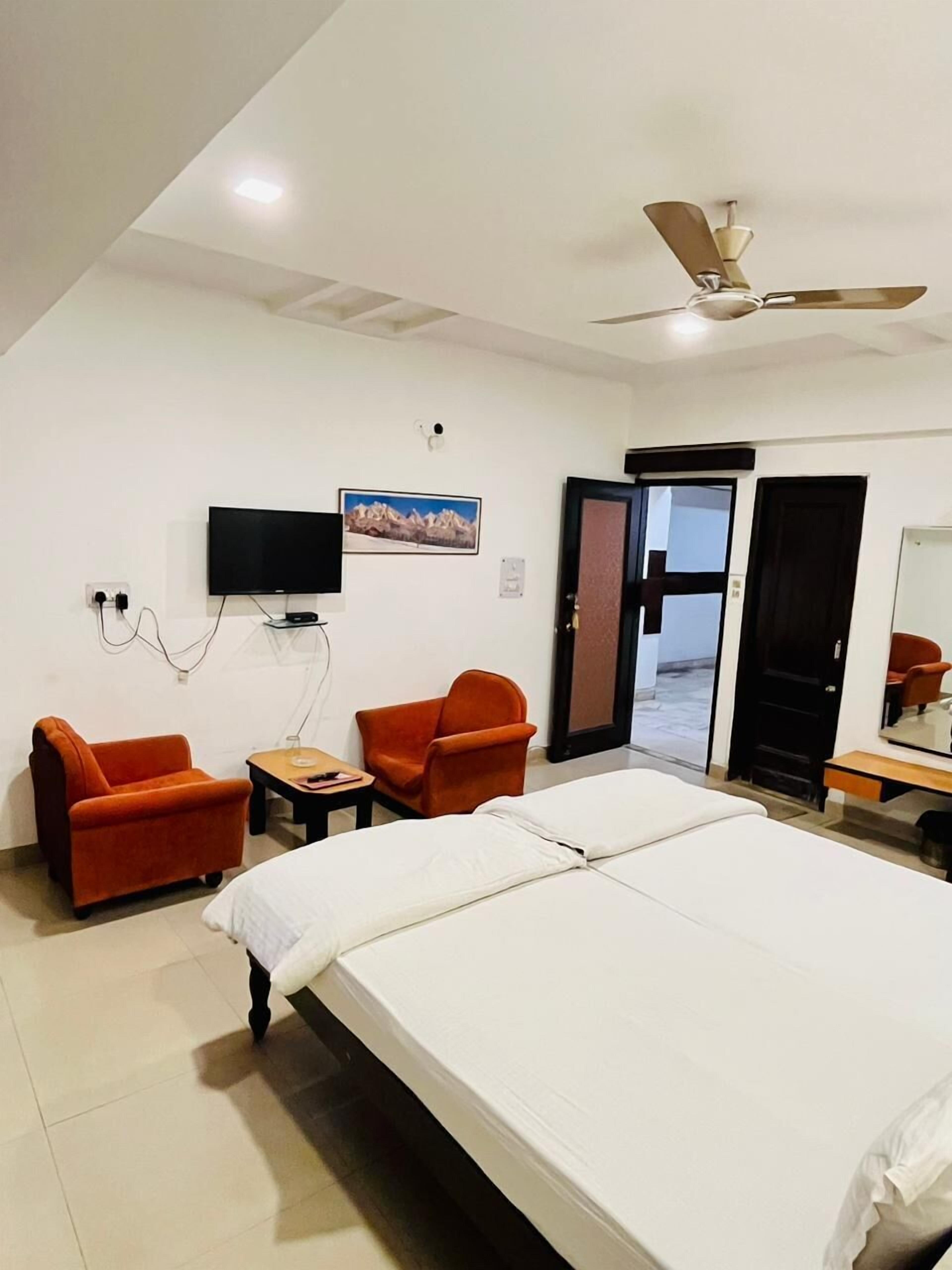 A hotel room with a bed, two orange armchairs, a small table, a television, and a ceiling fan.