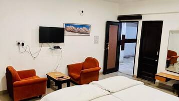 A hotel room with a bed, two orange armchairs, a small table, a television, and a ceiling fan.