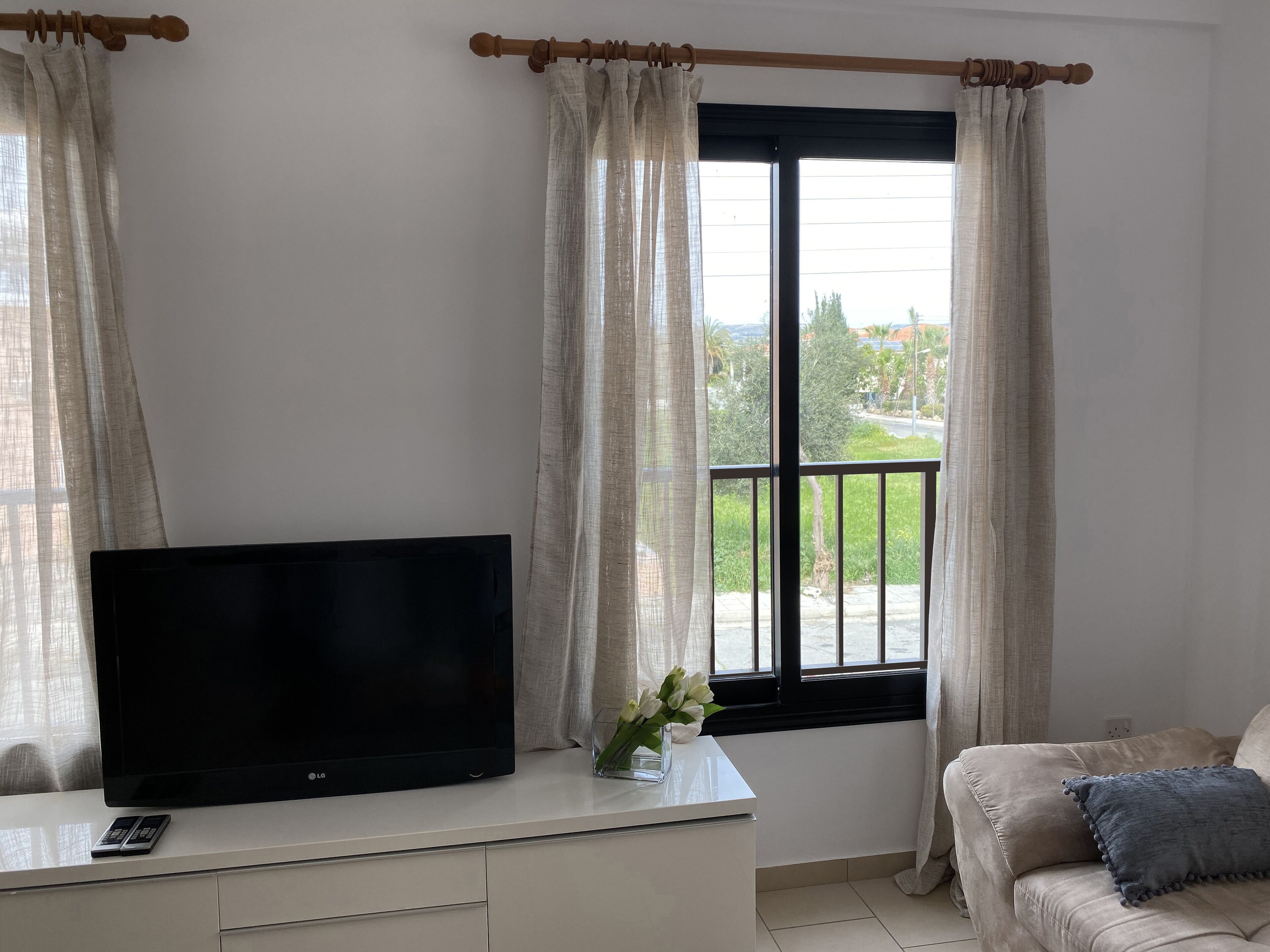 A living room with a flat-screen TV on a white cabinet, a remote control, a vase with flowers, and a beige sofa with a blue pillow.