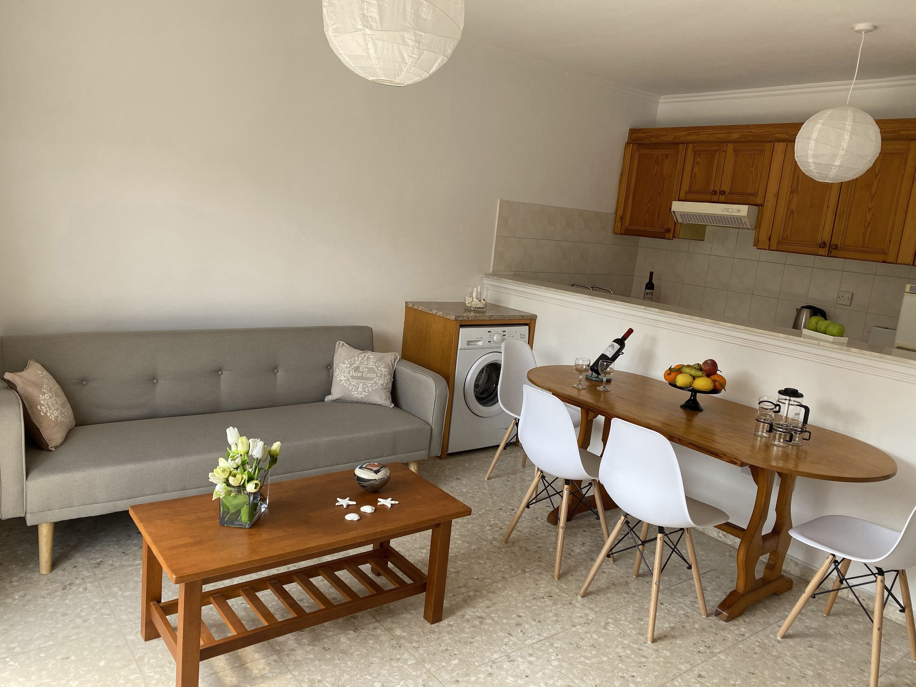 A modern living room with a grey sofa, wooden coffee table, and a dining area with a wooden table and white chairs. A washing machine is visible in the background.