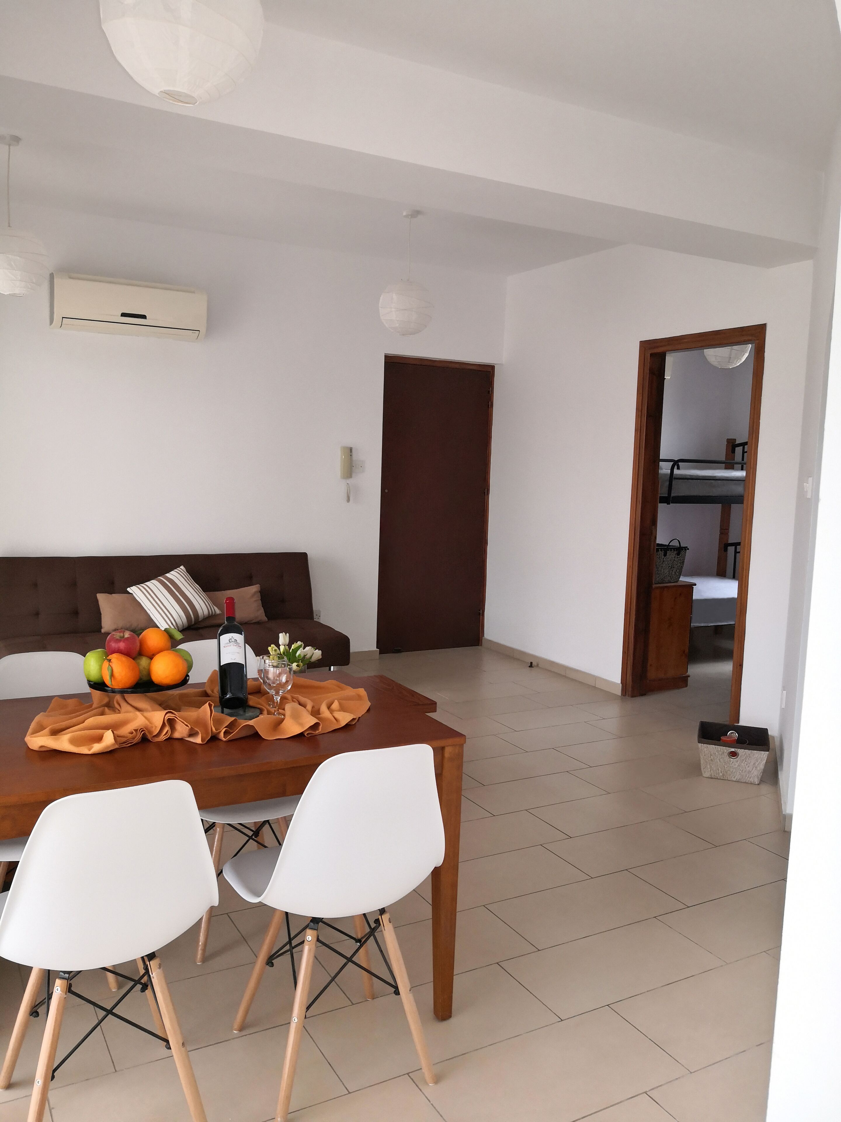 A dining area with a wooden table, two white chairs, a brown sofa, and a fruit bowl with oranges and apples.