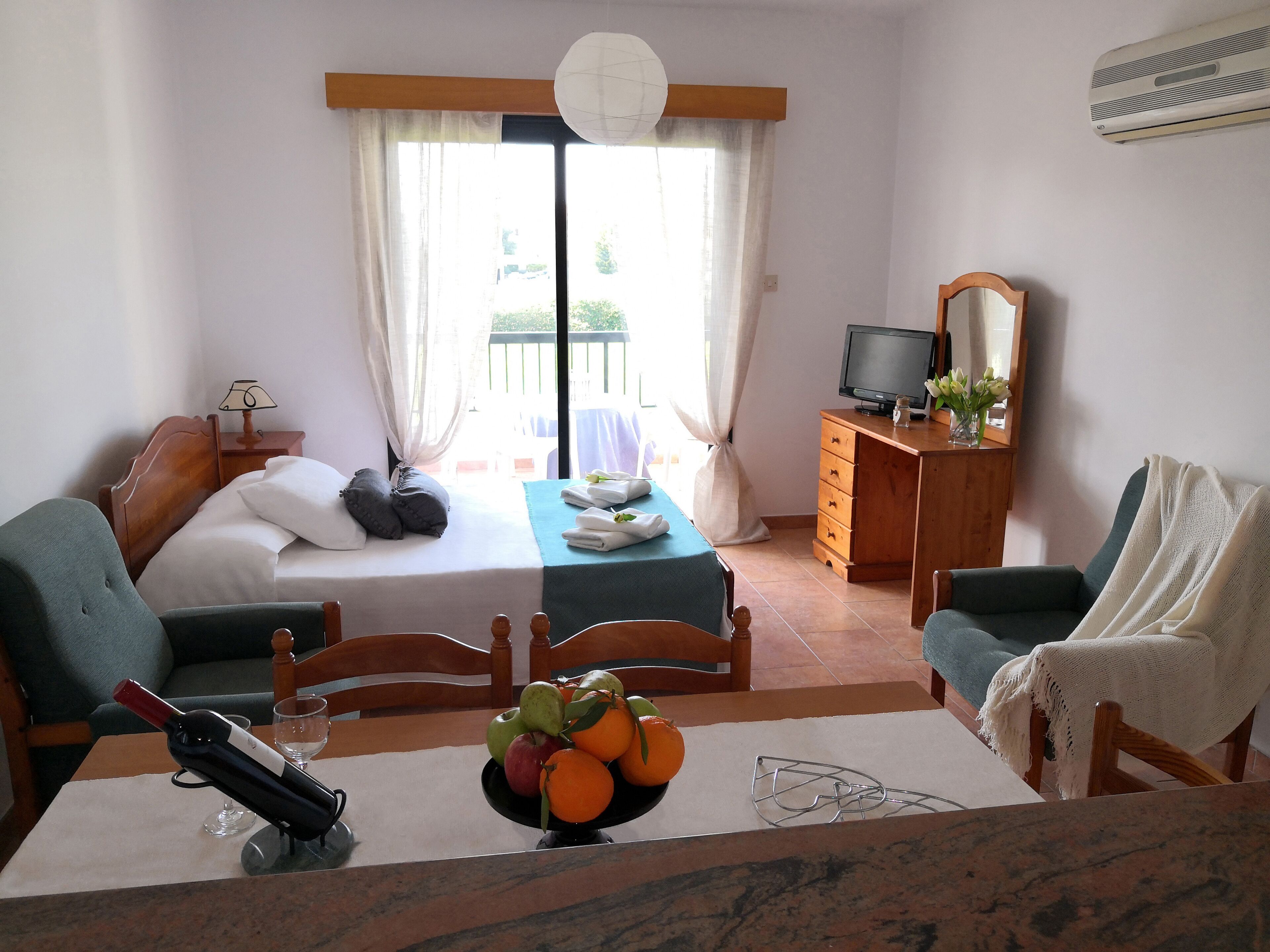 A hotel room with a bed, a desk with a computer, a dining table with fruit, and a balcony with a view.