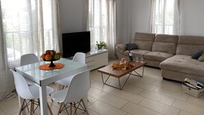 A living room with a beige sofa, a glass coffee table, a TV on a stand, and a dining table with chairs. - Seashell Apartments (Paphos)