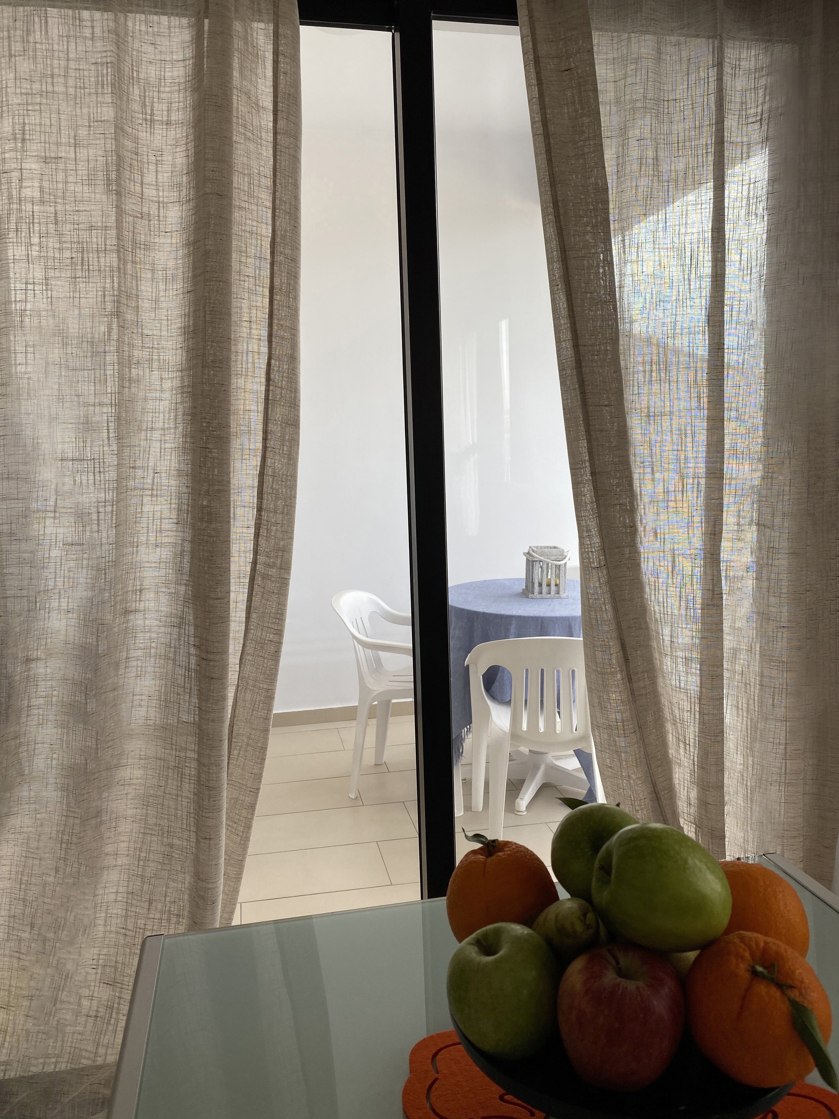 Une pièce avec une table, un saladier de fruits et une vue par la fenêtre, qui est ornée de rideaux.