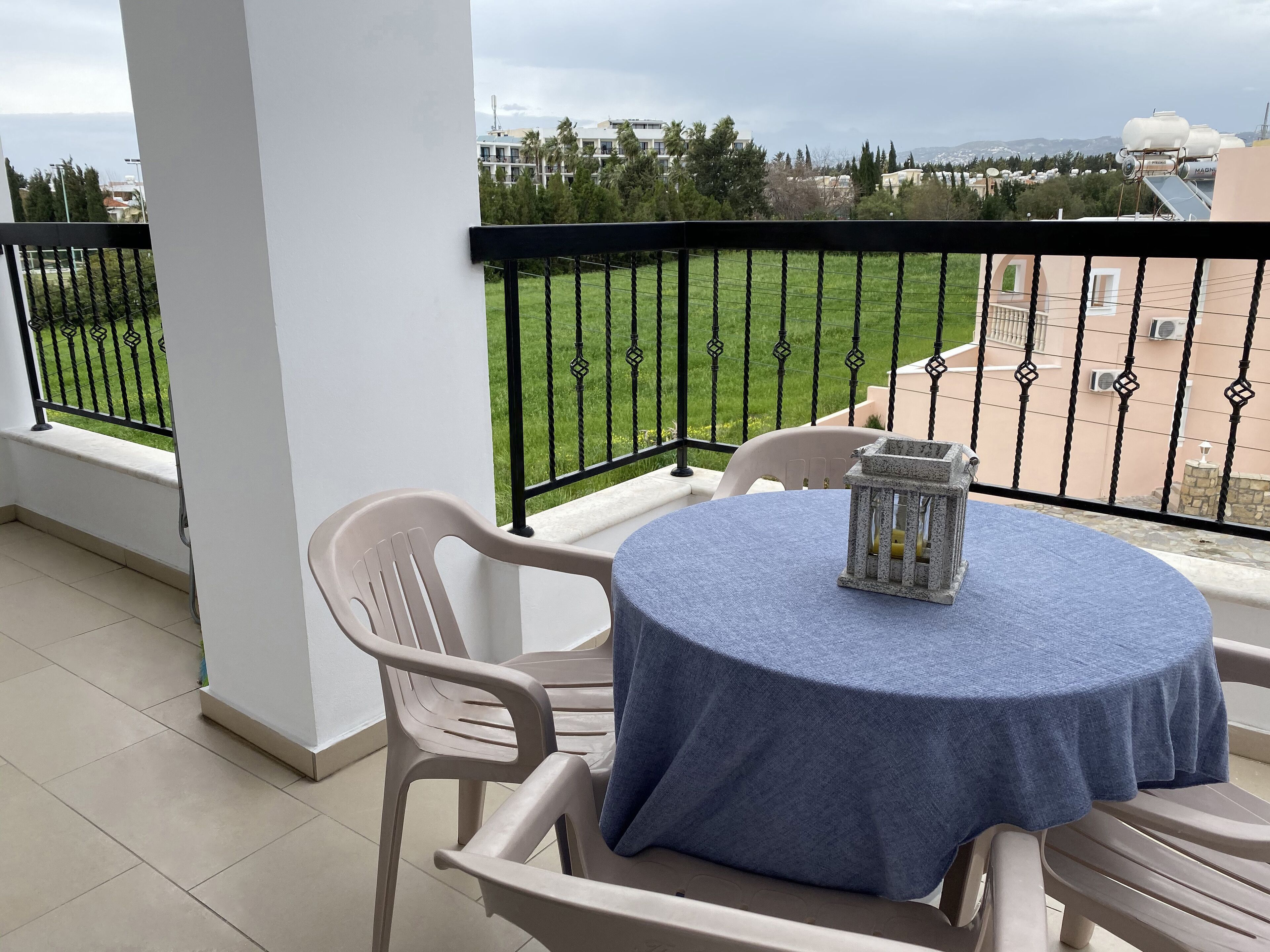 En balkon med et bord og stole, udsigt til en grøn græsplæne og bygninger i baggrunden.