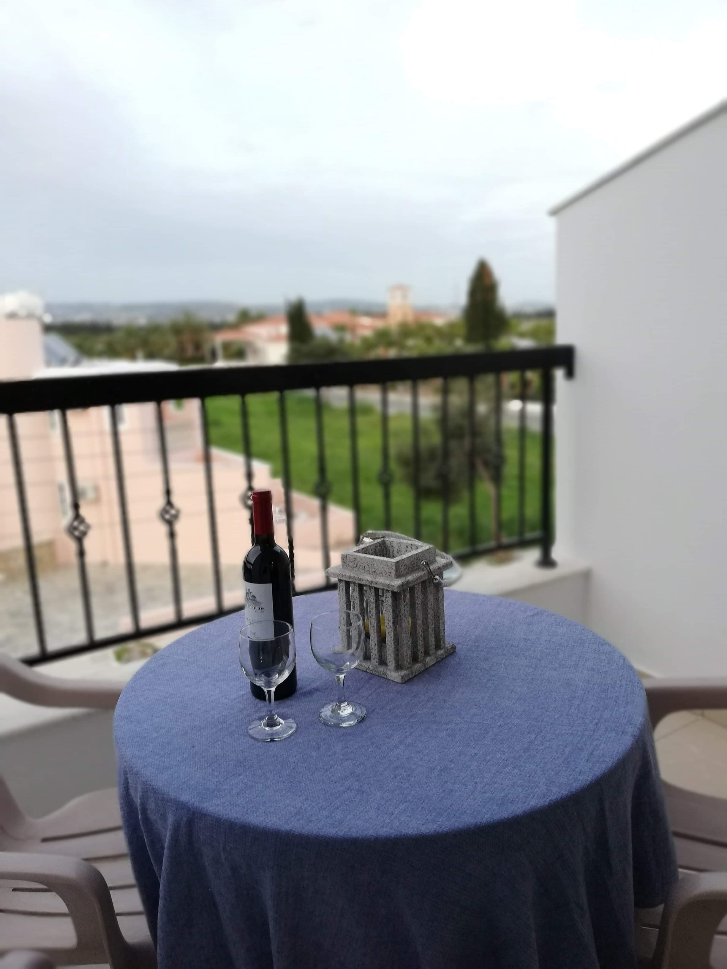 A round table with a blue tablecloth, two wine glasses, and a wine bottle on a balcony overlooking a cityscape.