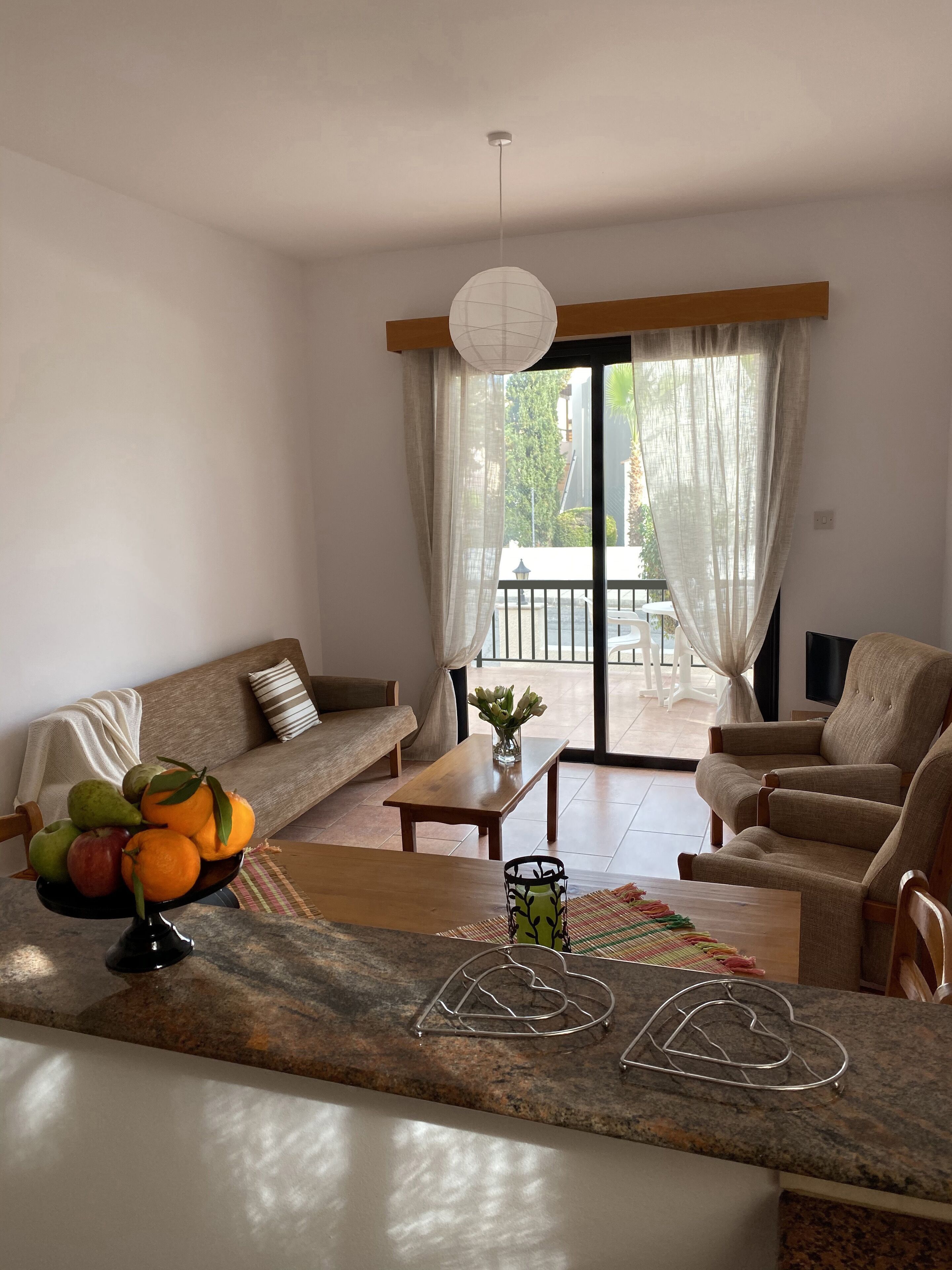 Un salon moderne avec un canapé, un fauteuil et une table à manger. Un panier de fruits est posé sur la table, et on peut voir le paysage extérieur à travers la fenêtre.