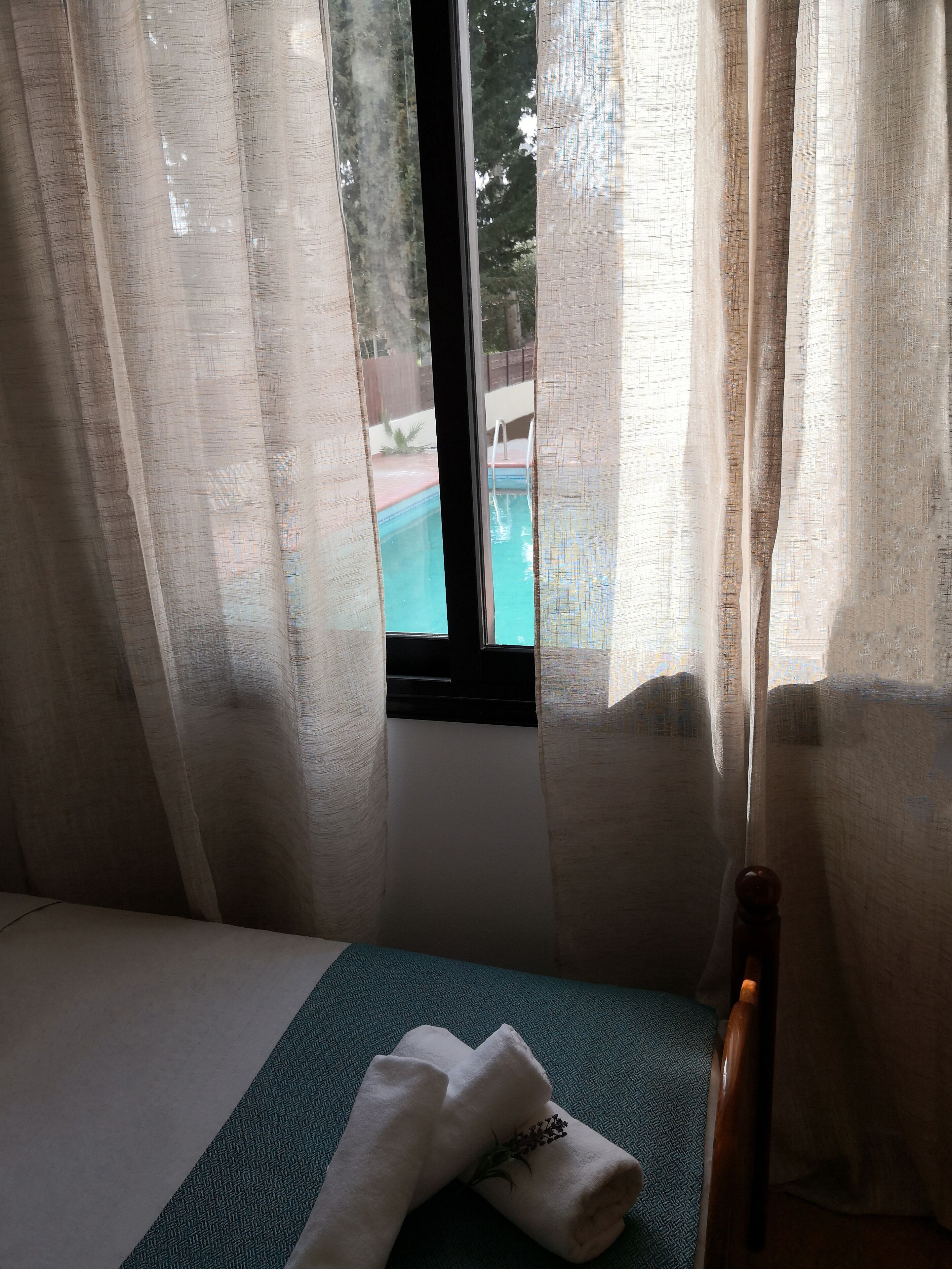 A room with a bed, a window with sheer curtains, and a view of an outdoor swimming pool.