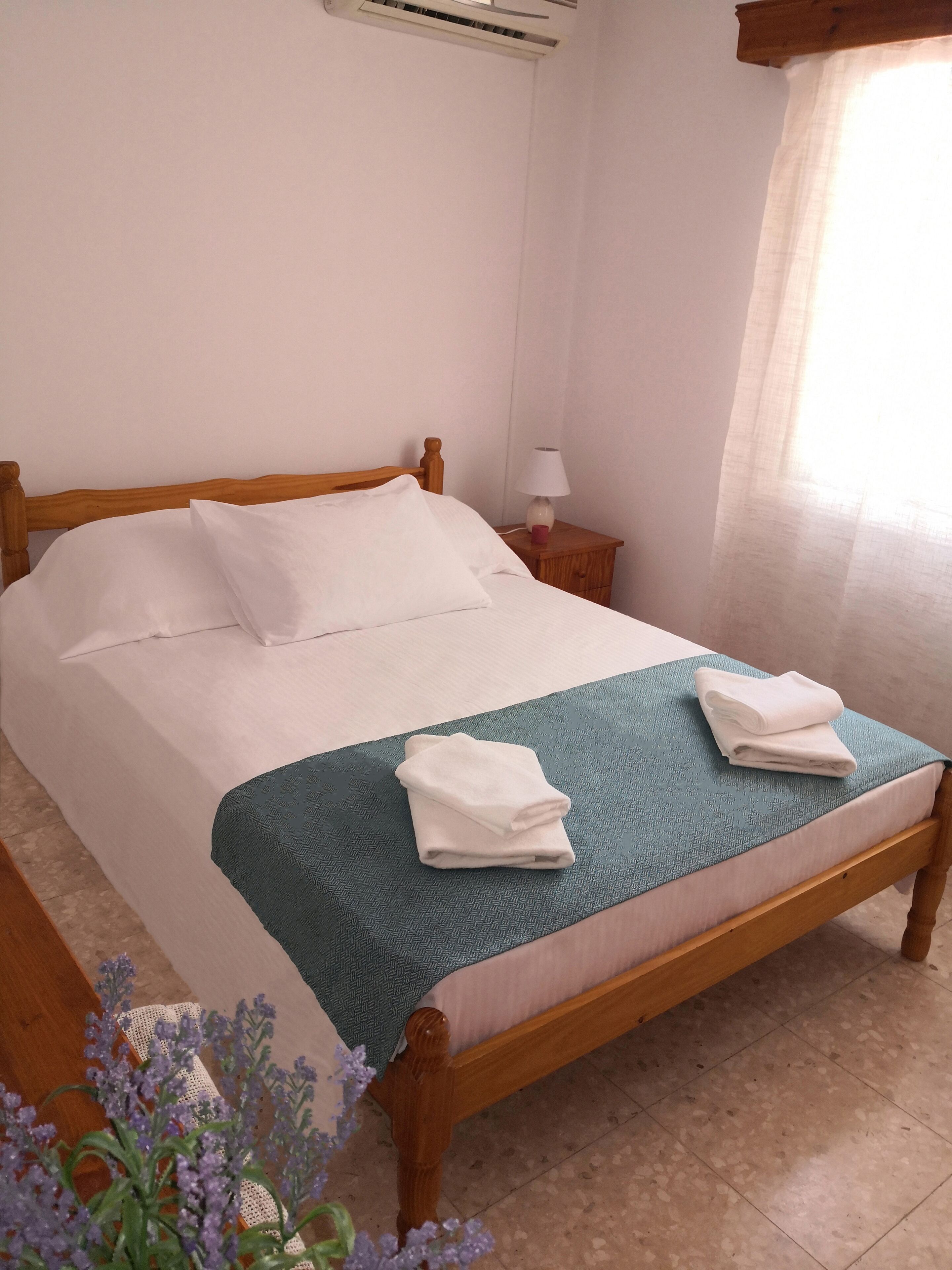 Una cama bien hecha con sábanas blancas, una manta color turquesa y toallas dobladas sobre un cabecero de madera.