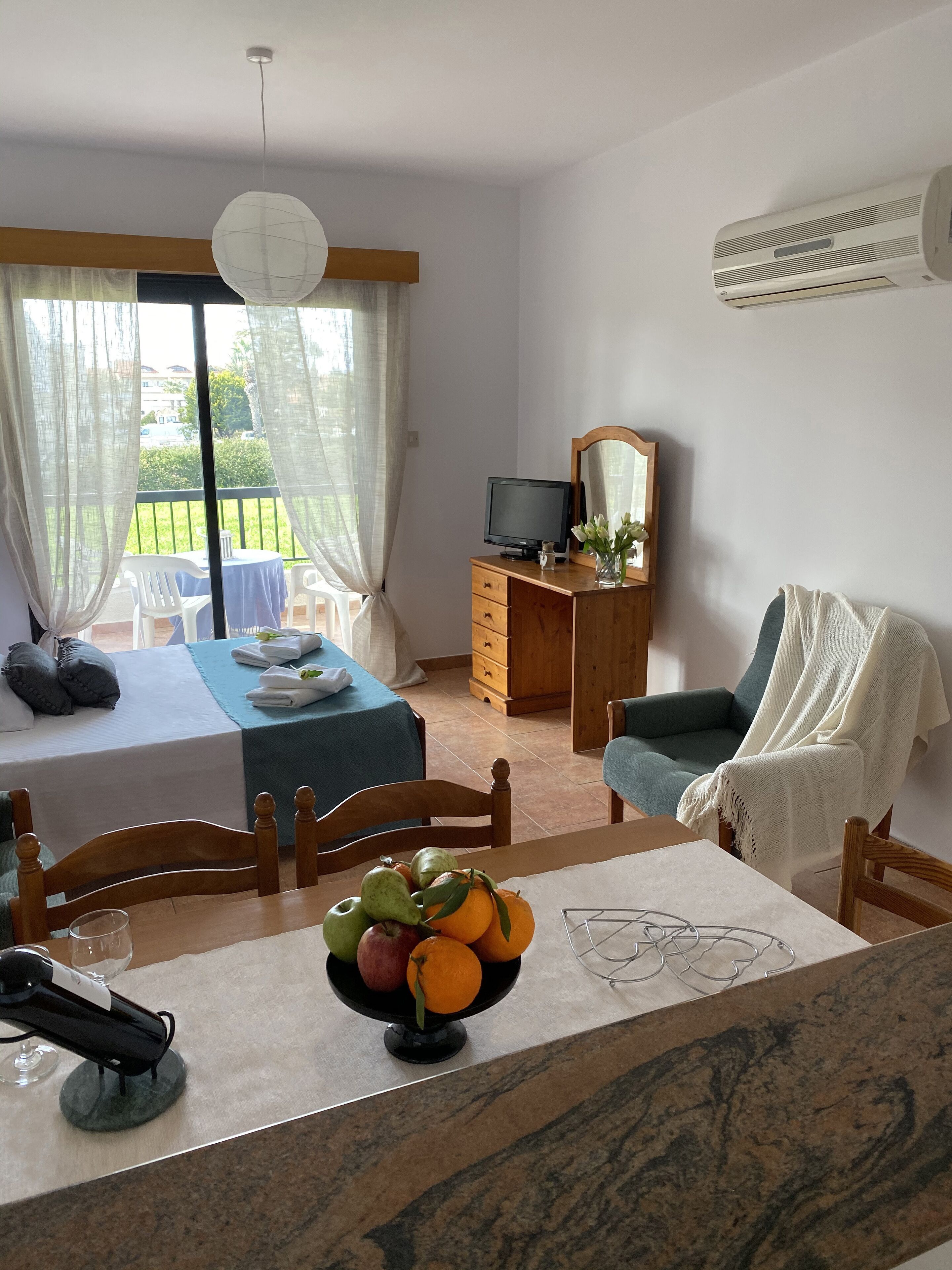 A room with a dining table, chairs, a fruit bowl, a TV, a mirror, and a window with curtains.