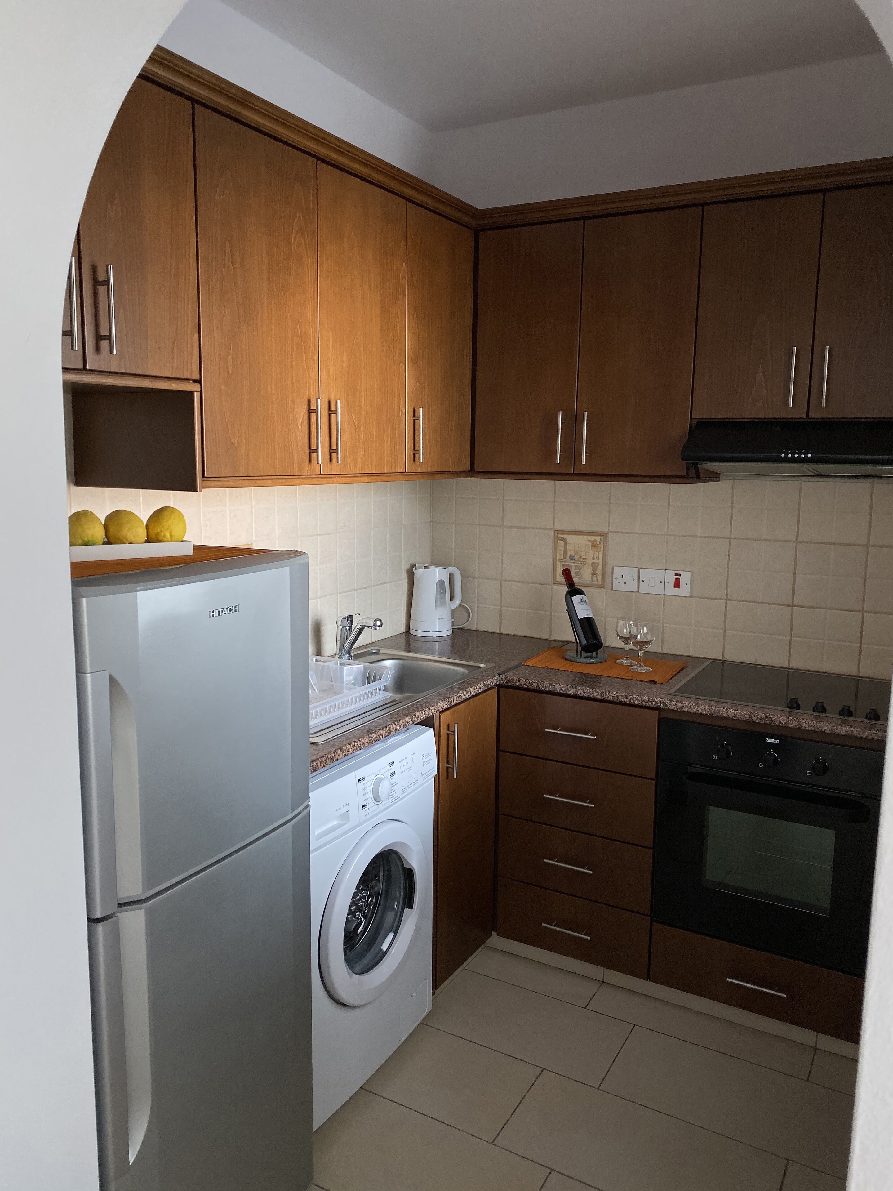 Cocina compacta con armarios de madera, un refrigerador de acero inoxidable, lavadora, cafetera y estufa.