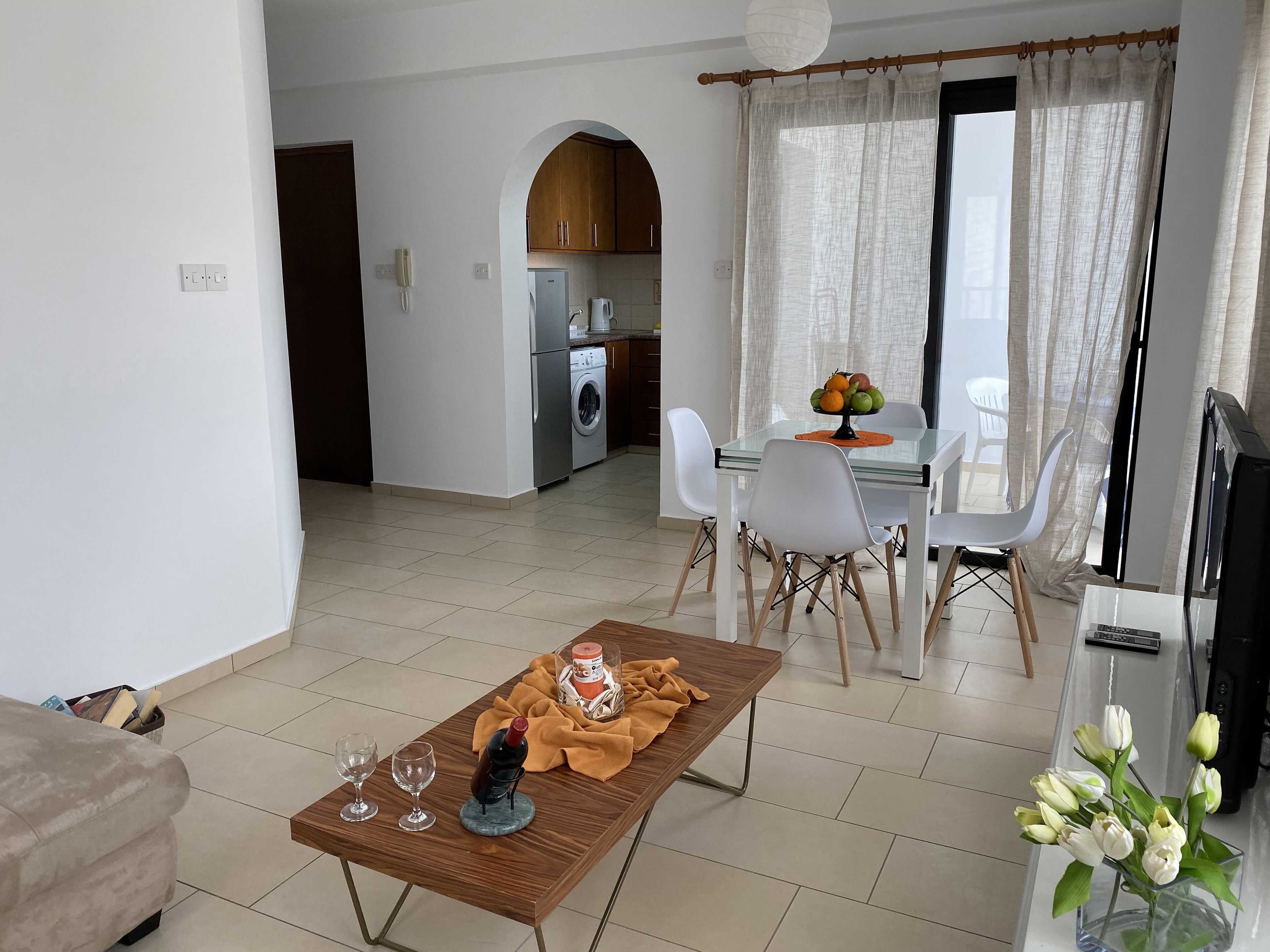 A modern living room with a dining area, a kitchen in the background, and a view through a window with curtains.