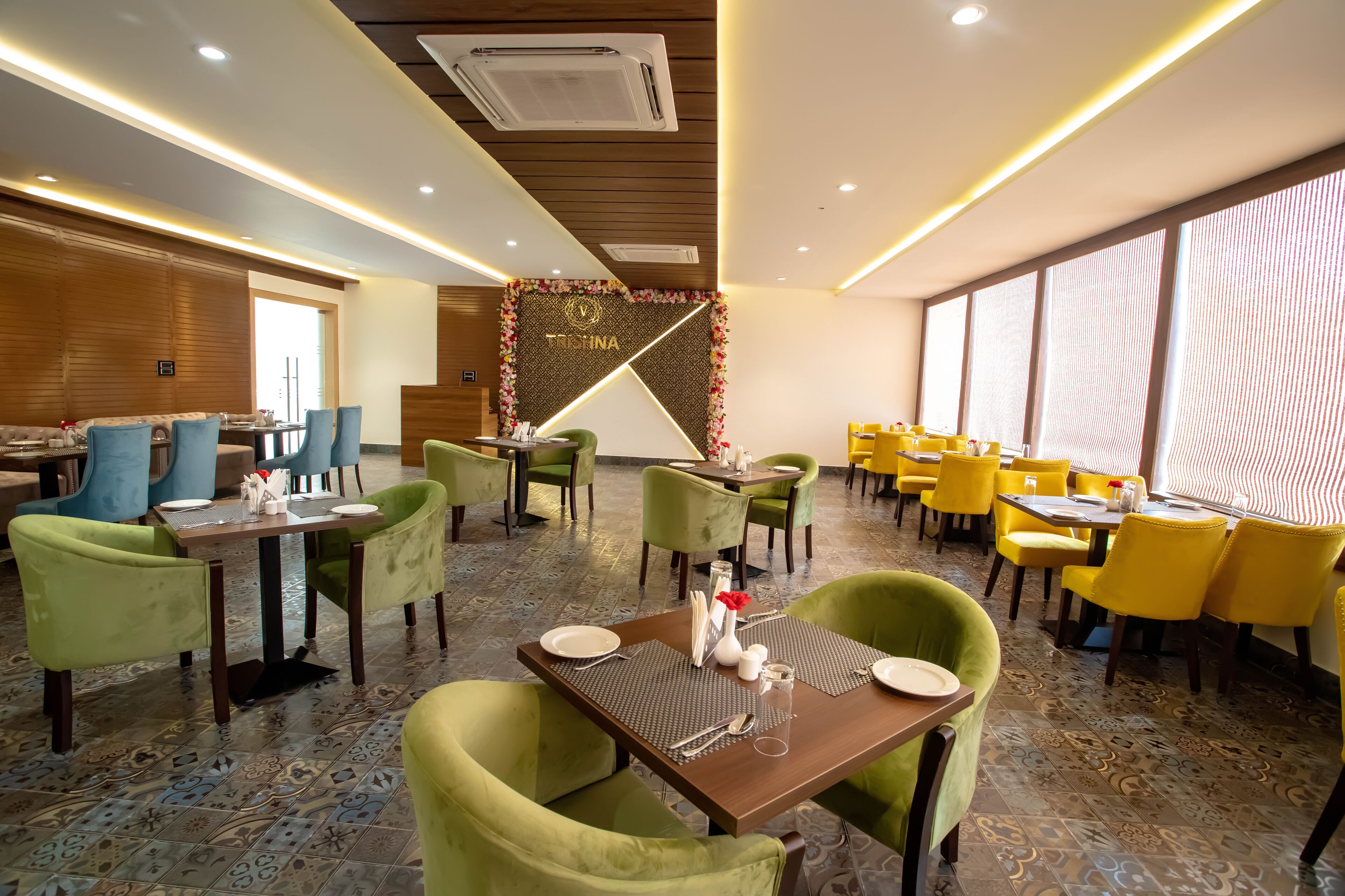 Un restaurante con sillas verdes y amarillas, mesas de madera y una pared decorativa con un diseño geométrico.