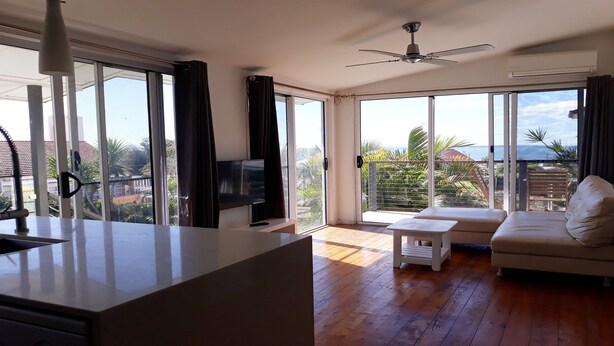 Een moderne woonkamer met een televisie, een bank, een klein tafeltje en grote schuifdeuren die uitkomen op een balkon met uitzicht op zee.