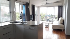Een moderne keuken met een vaatwasser, een spoelbak en een raam met jaloezieën. Er is een balkon met een witte bank en een klein tafeltje.