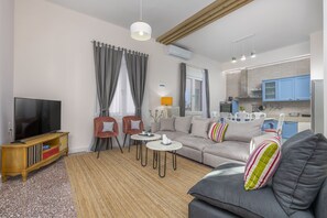 Un salon moderne comprenant un canapé, deux fauteuils, une table basse, un téléviseur et un coin cuisine avec des meubles bleus et un plan de travail.