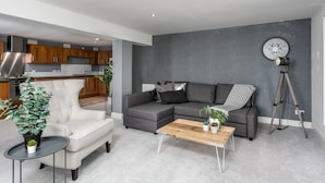 Un saló modern amb un sofà seccional gris, una taula de centre rodona petita i un butaquesa gris. Hi ha una làmpada de trípode i una planta en test amplesa al sòl. La habitació té una paret gris amb textura i una zona de cuina visible amb armaris de fusta i una campana extractora d'acer inoxidable.
