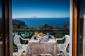 A balcony table with a patterned tablecloth, white plastic chairs, and a breakfast setting with pastries, a jug of orange juice, and a coffee pot.